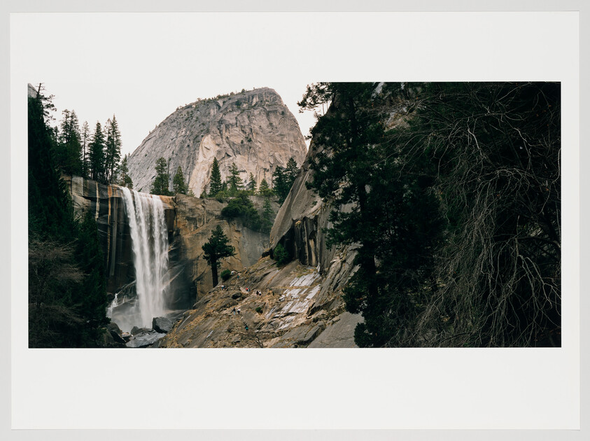 A scenic view of a waterfall cascading down a sheer rock face into a pool below, with a backdrop of a large granite cliff and surrounded by pine trees. Some visitors can be seen at a distance on the rocky terrain near the waterfall base.