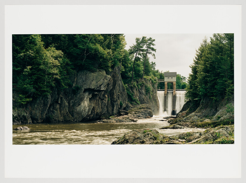 A scenic view of a waterfall cascading down from a small dam with a building on top, surrounded by lush green trees and rocky cliffs. The turbulent water flows through a rugged landscape, indicating a natural river setting.