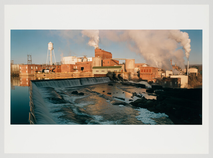 A sprawling industrial complex with multiple red brick buildings beside a waterfall. A water tower stands in the background, and plumes of steam or smoke rise from tall stacks into the clear sky. Reflections of the buildings and the waterfall are visible on the calm water surface in the foreground.