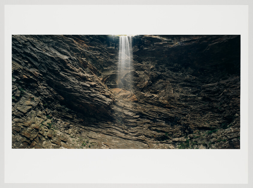 A slender waterfall cascades down a rugged cliff face, with sunlight filtering through the mist at the top. The rocky surface surrounding the waterfall is layered and textured, with sparse vegetation clinging to the crevices.