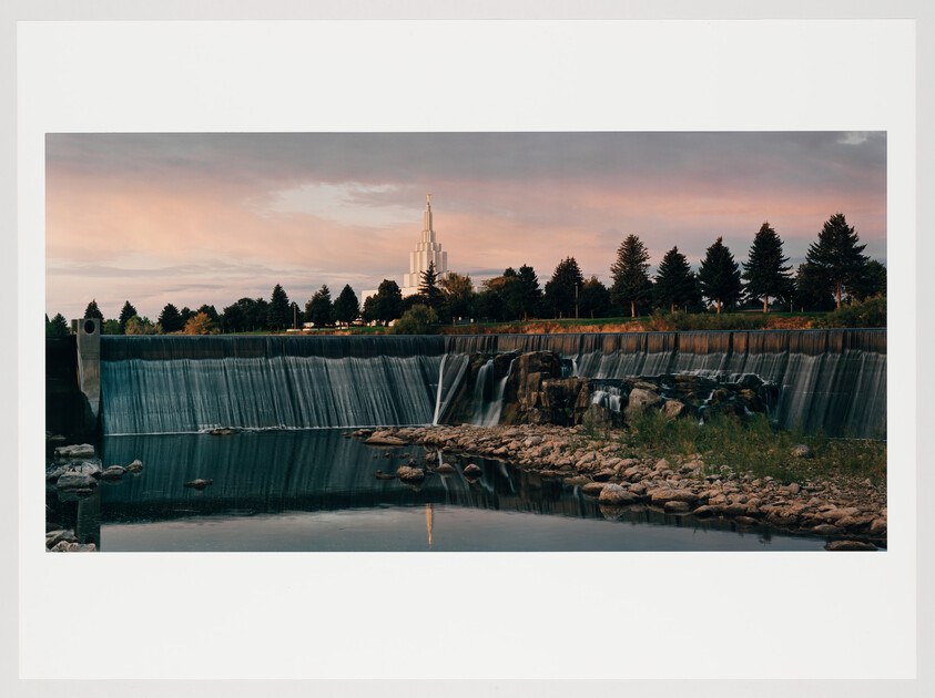 A serene waterfall cascades over a wide ledge into a calm river at dusk, with a spire-topped building silhouetted against a pastel-hued sky in the background.