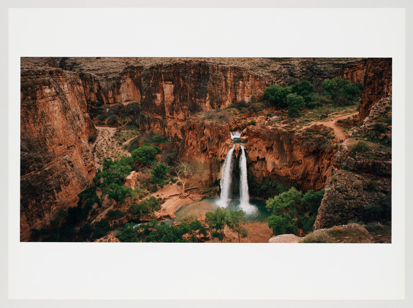 A panoramic view of a majestic waterfall cascading down a red rock cliff into a serene pool below, surrounded by green vegetation and set against a backdrop of a rugged canyon landscape.