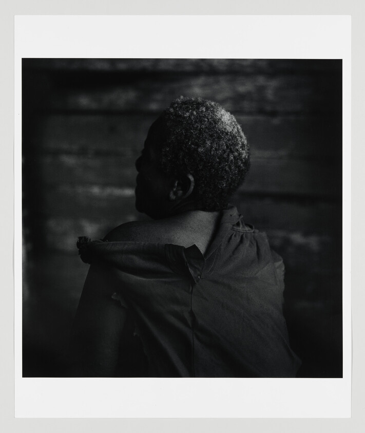 Older person with short gray hair turned away, wearing a torn shirt against a dark wooden wall.