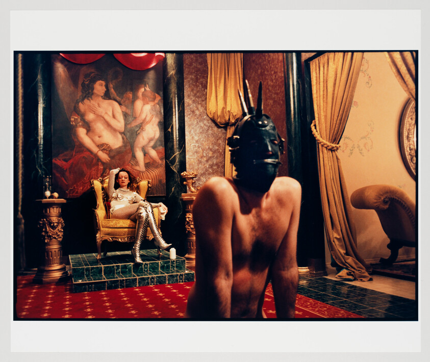 A masked, shirtless person kneels in front of a woman lounging on a gold throne in a lavish room.