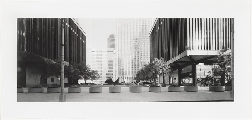 Wide urban plaza between tall office buildings with trees, planters, and a central sculpture.
