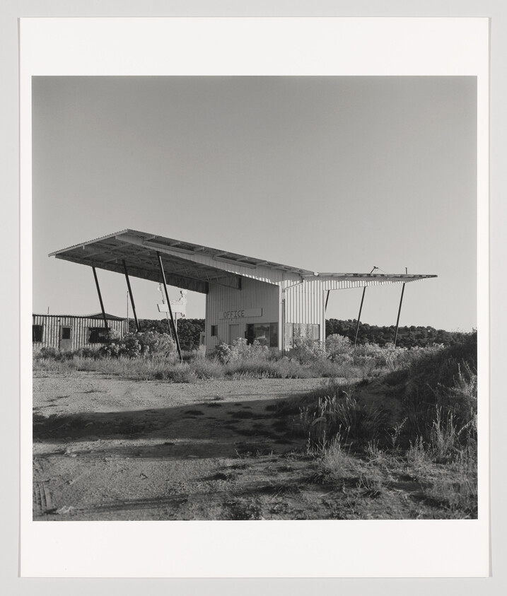 Abandoned roadside office building with a large slanted canopy and overgrown weeds.