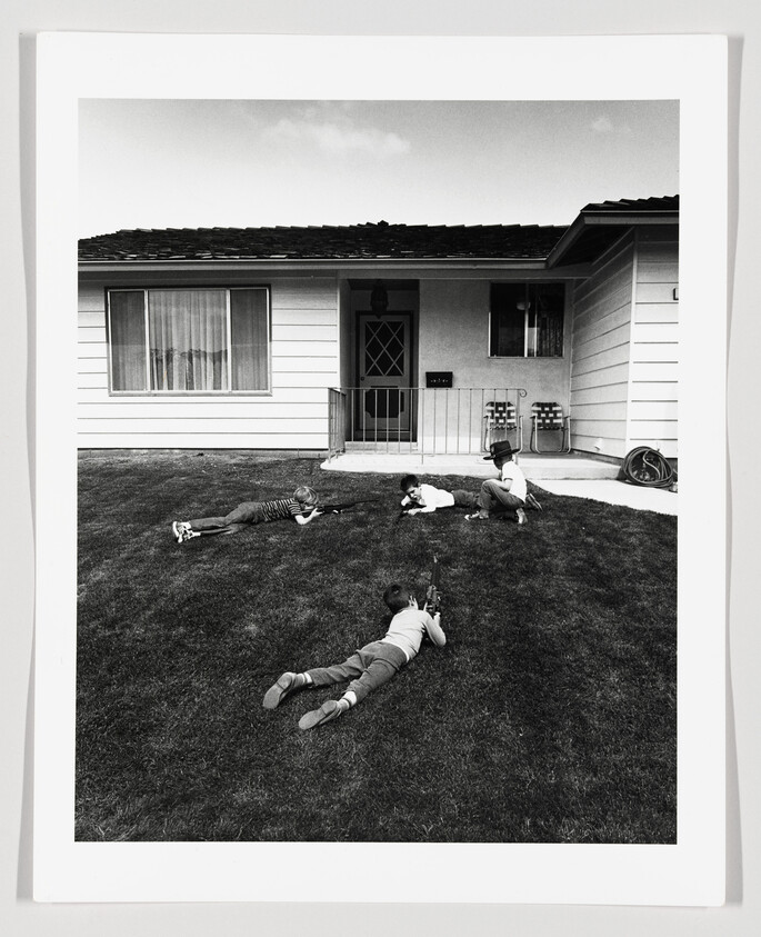 Four children lie on a front lawn pretending to aim toy rifles toward the house.