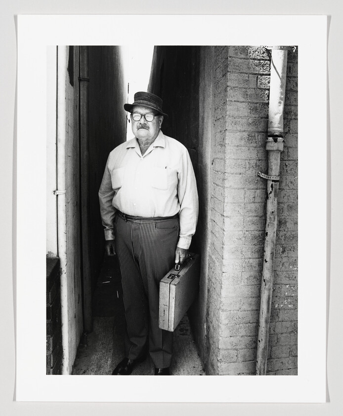 A black and white photograph of an older man standing in a narrow alleyway. He is wearing glasses, a hat, a light-colored shirt, and striped trousers. He holds a briefcase in his right hand and has a stern expression on his face. The alley walls are textured, and there's a pipe running down the wall to his right.