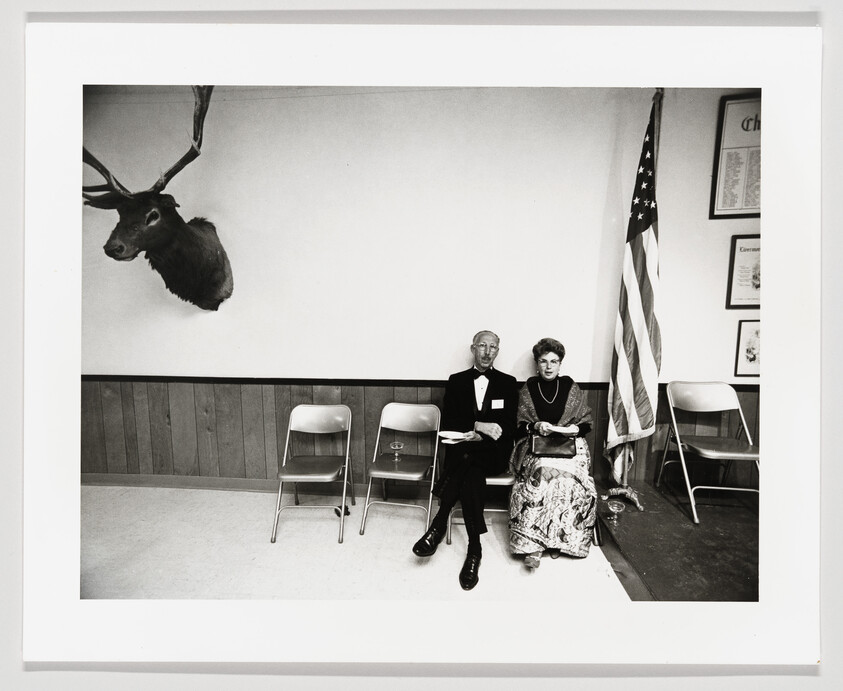 An elderly couple in formal dress sit on folding chairs beside an American flag and a mounted deer head.