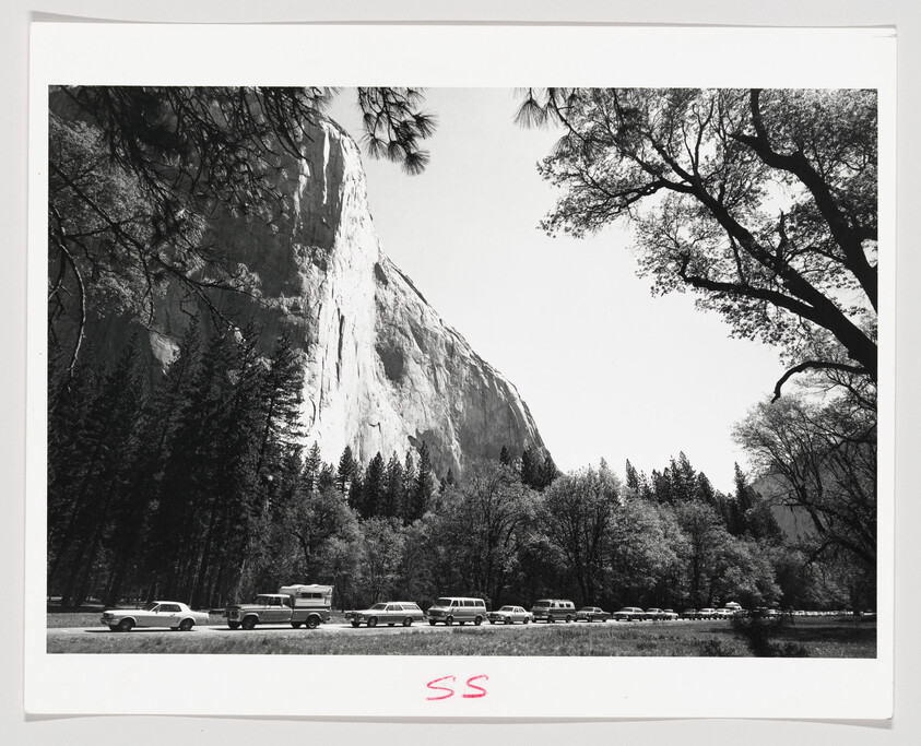 A black and white photograph depicting a majestic cliff face towering over a forested area with a line of vintage cars parked at the base. The trees in the foreground frame the rock formation, and there is a clear sky above. The image has a white border with "SS" written in red in the bottom right corner.