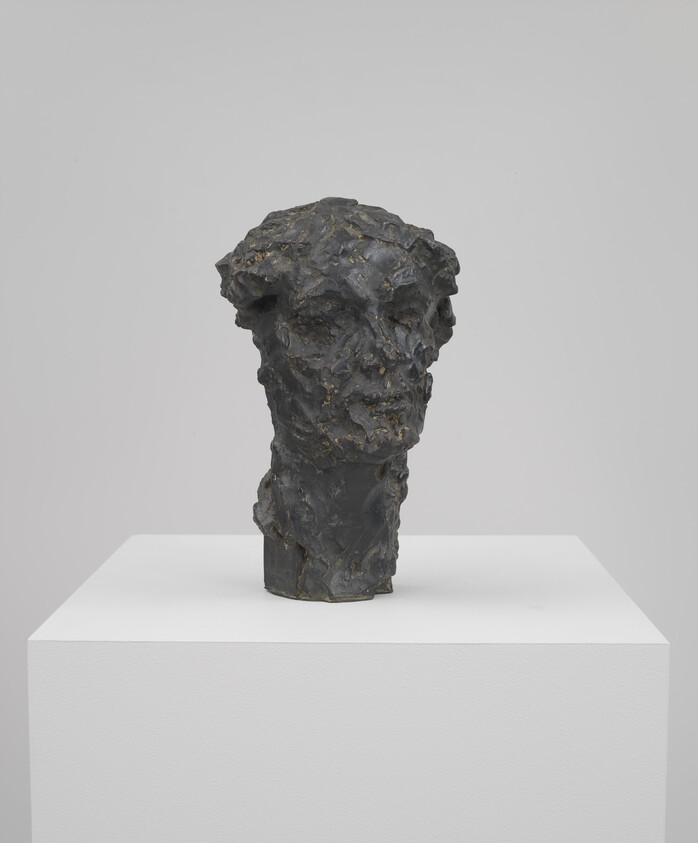 Rough-textured dark bronze sculpture of a human head displayed on a white pedestal.