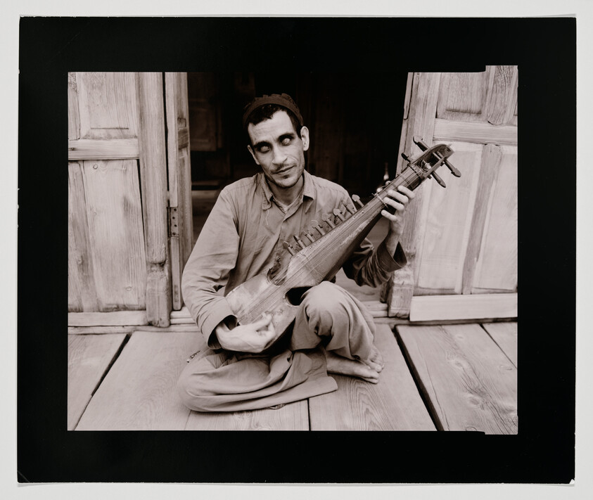A seated man plays a traditional stringed instrument while sitting in a wooden doorway.