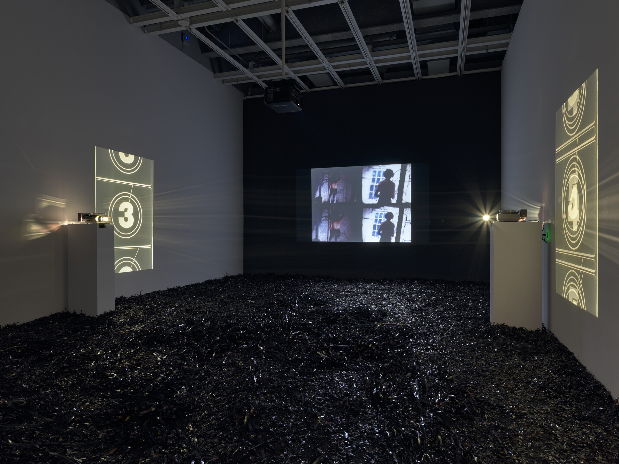 Dark room with film projectors displaying countdowns and silhouettes on walls, floor covered in black film strips.