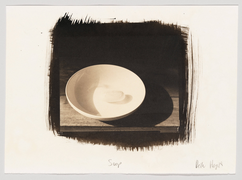 A sepia-toned artwork featuring a bowl on a wooden surface with a dark, roughly painted border. The bowl casts a shadow on the surface, and there are handwritten inscriptions at the bottom, possibly the title "Soup" and the artist's signature.