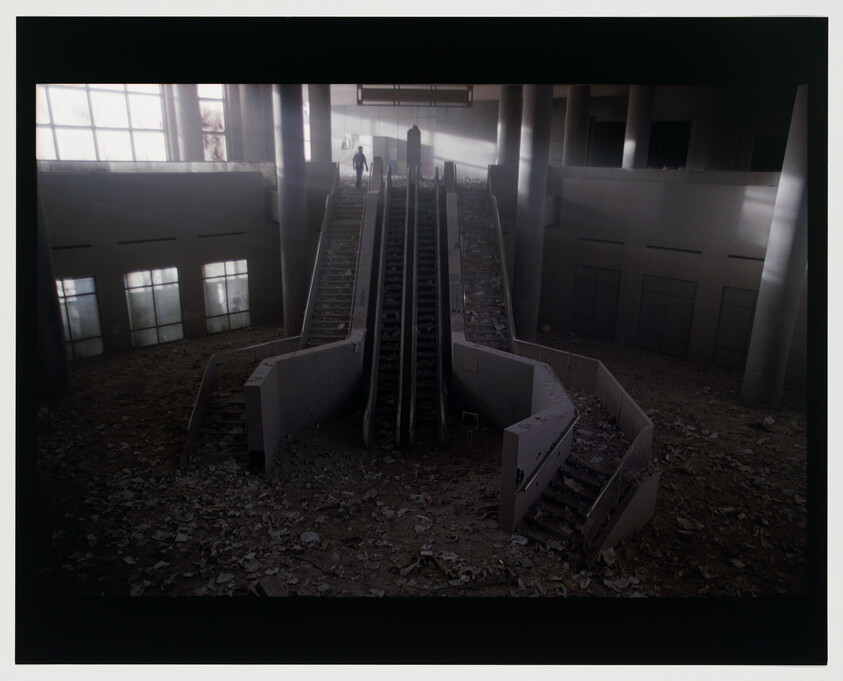 Abandoned, debris-covered escalators rise in a dark, empty atrium with a lone figure at the top.