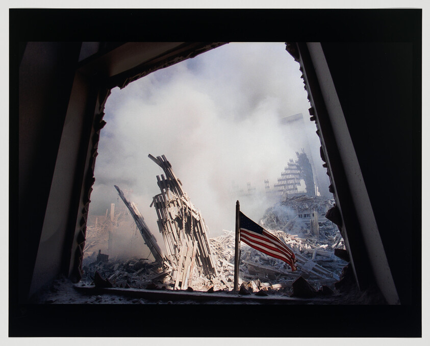 American flag standing amid smoke and twisted steel debris at a destroyed building site.