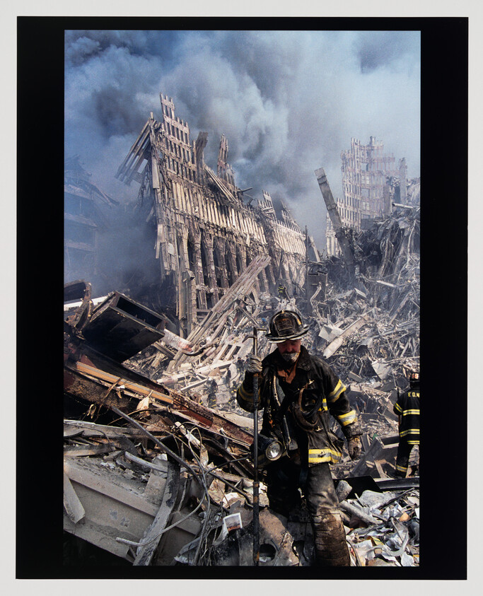 A firefighter walks through smoldering rubble with collapsed steel towers and heavy smoke in the background.