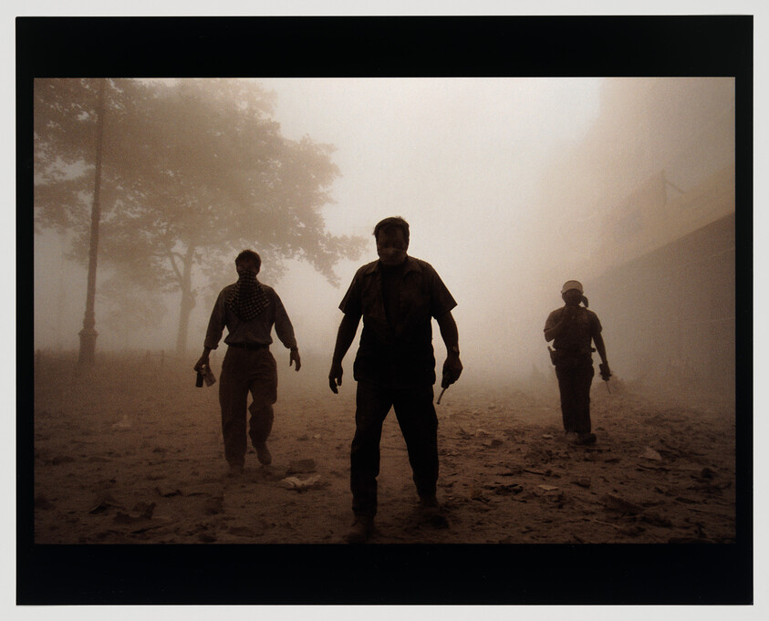 Three silhouetted figures walk through thick dust and debris, one holding a camera and another a radio.