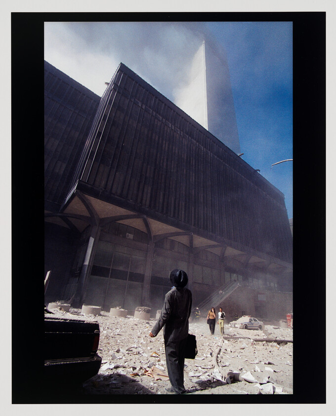 A suited person stands amid rubble and dust, looking up at a damaged high-rise while others walk nearby.