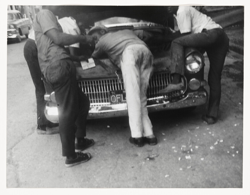 Three men lean into the open hood of an old car, inspecting or repairing the engine.