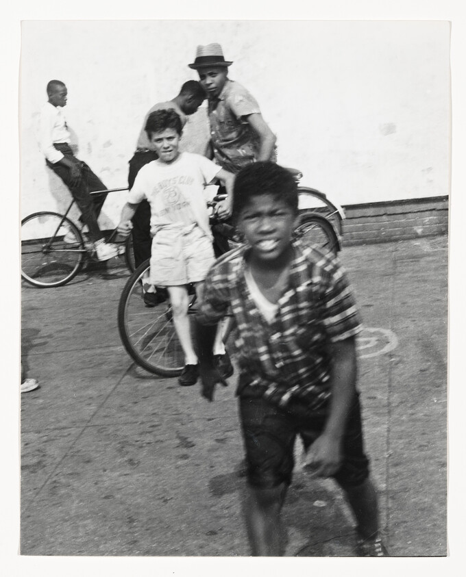 A boy runs toward the camera while other boys stand with bicycles against a wall.