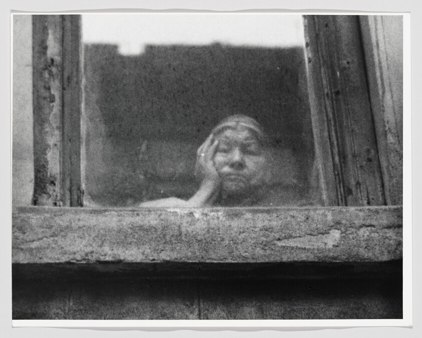 An elderly woman rests her head on her hand while gazing out a grimy window.