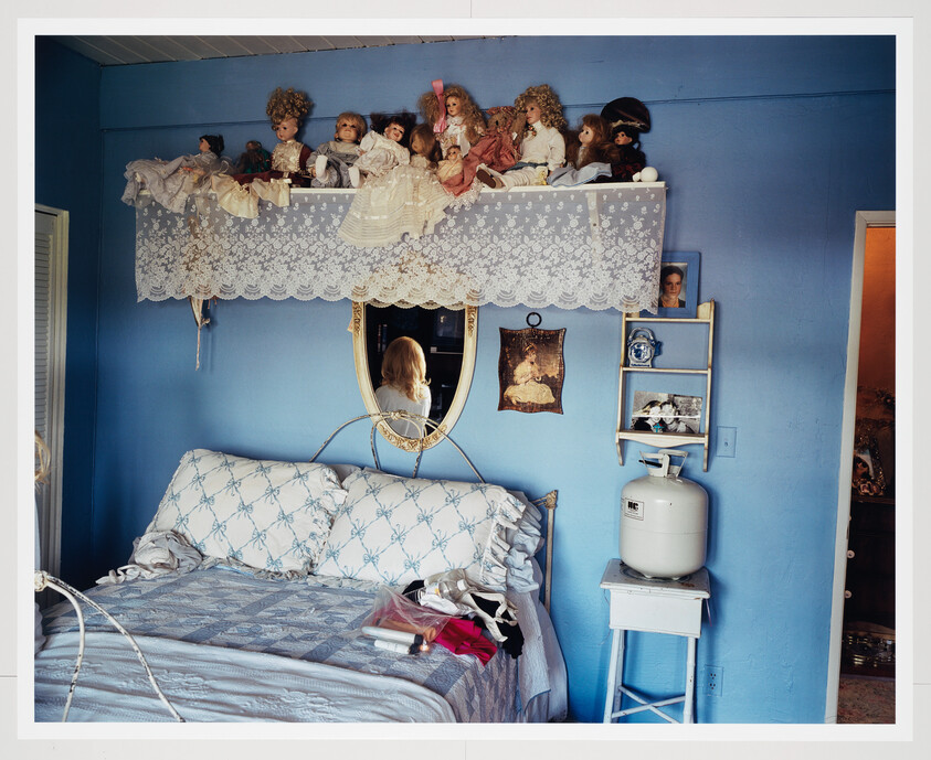 Blue bedroom with a row of porcelain dolls on a lace shelf above a bed and mirror.