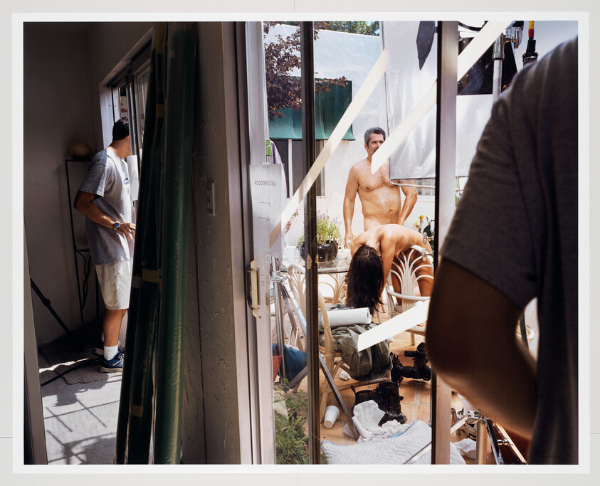 Two people pose nude on a set while crew members adjust lighting and equipment nearby.