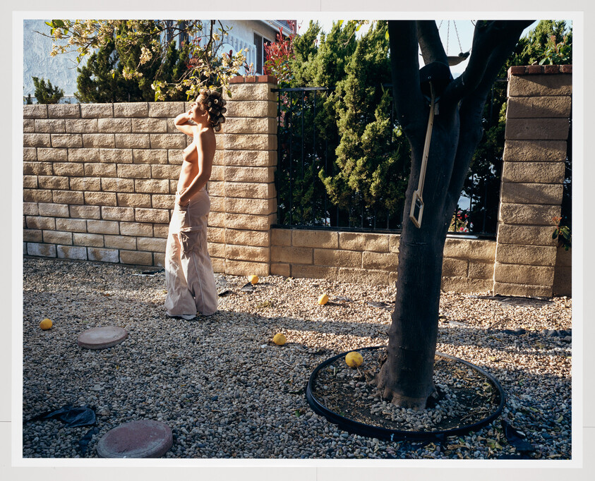 Person standing in a sunlit gravel yard with hair rollers, wearing loose pants and hand raised to forehead.