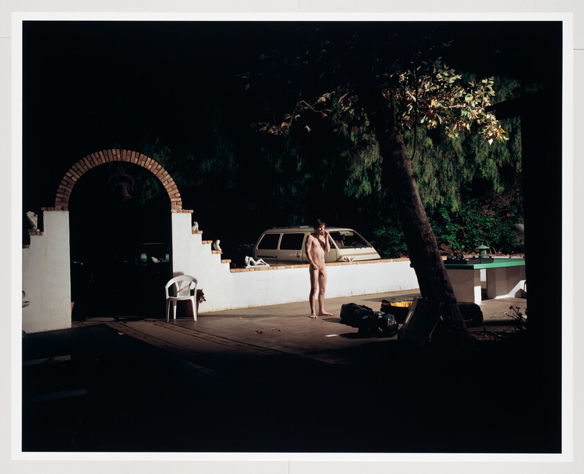 A naked person stands near a low white wall at night while holding a phone.