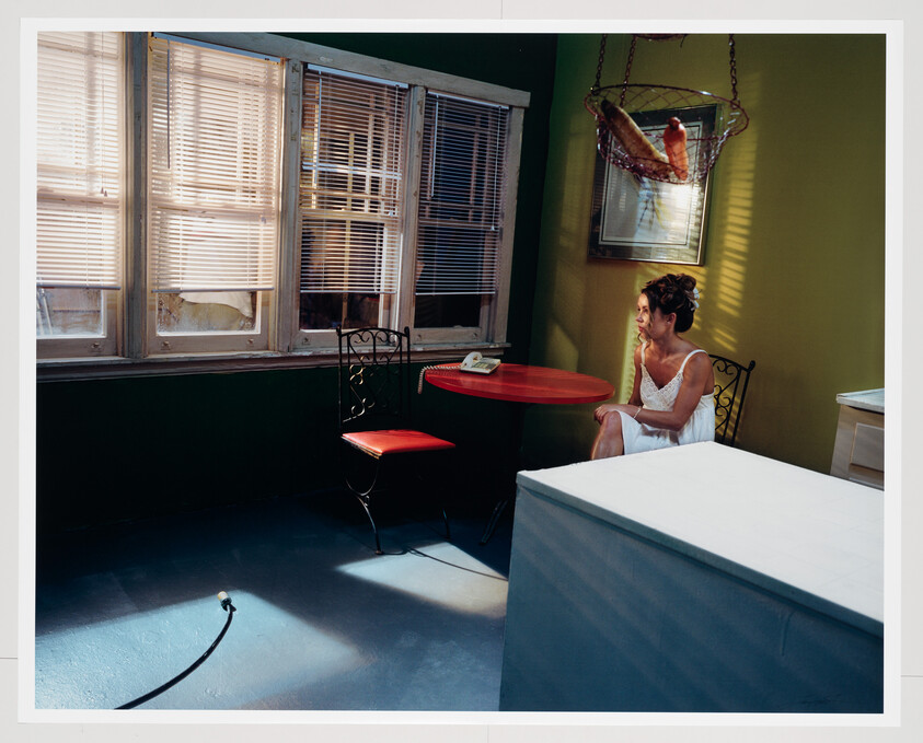 A woman in a white dress sits at a small red table by sunlit blinds, staring thoughtfully.