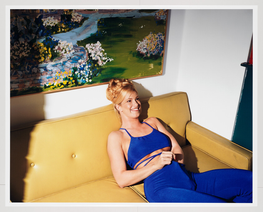 A smiling woman in a blue outfit reclining on a yellow couch under a painting.