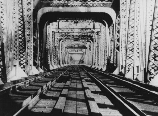 Railroad tracks run through a long steel truss bridge with repeating rectangular frames.