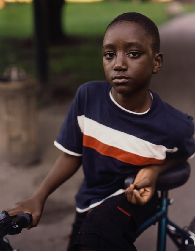 A young Black boy leans on a bike in the park and looks directly at the camera.
