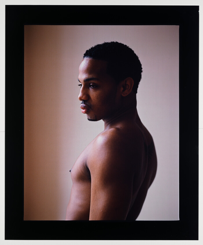 A profile portrait of a shirtless man looking to the side against a beige background, framed with a black border. The man has short hair and a thoughtful expression.