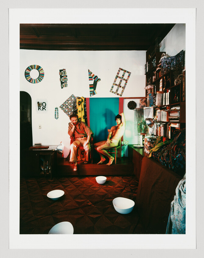 Two people sit in a colorful living room surrounded by books, wall art, and white bowls on floor.