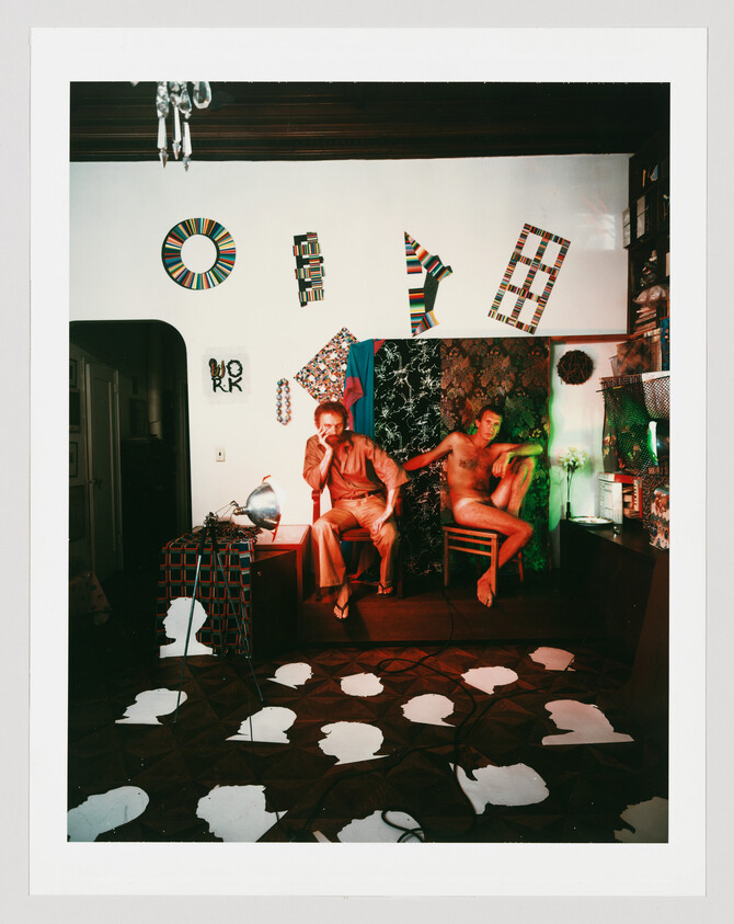 Two men pose in a colorful studio room, one fully clothed and one nude, surrounded by artwork.