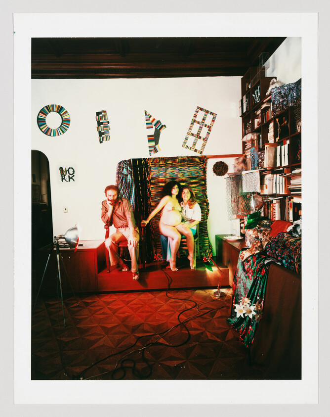 Three people pose in a cluttered, colorful room with a pregnant woman seated in the center.