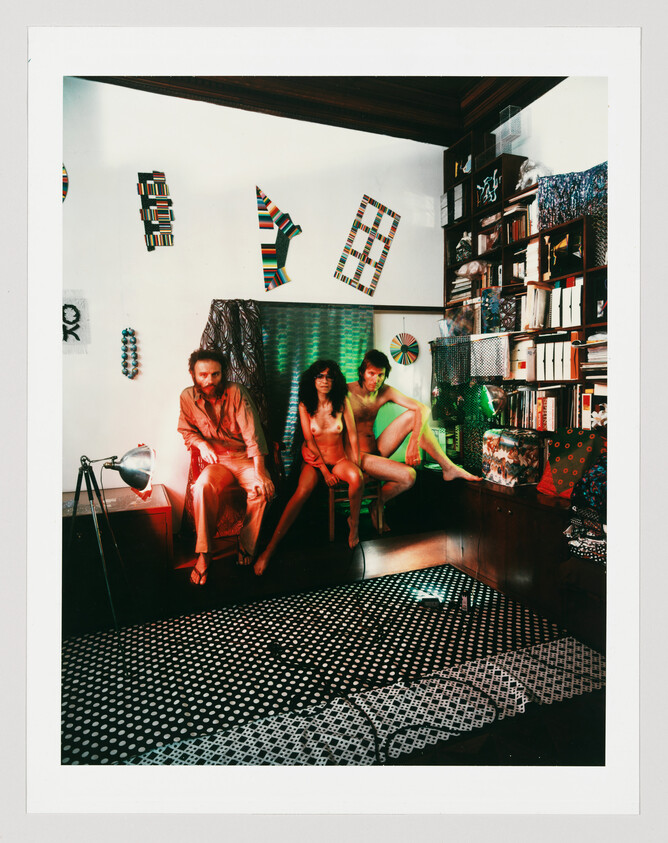 Three people sit closely on a bench in a colorful, patterned living room with bookshelves.
