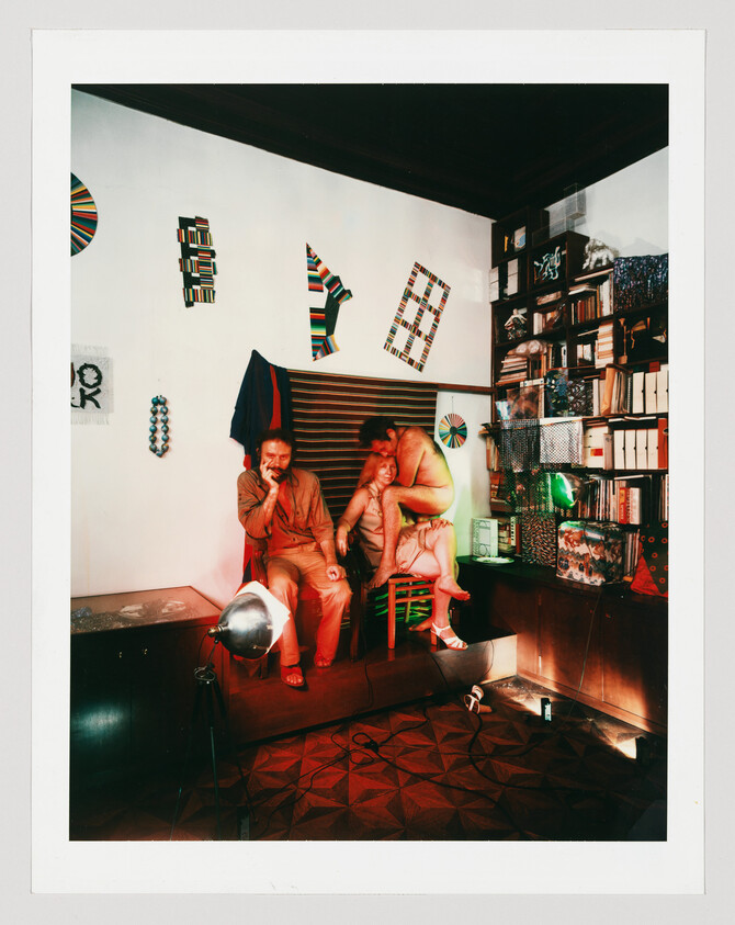 Three people in a cluttered living room sit and embrace amid bookshelves and colorful wall art.