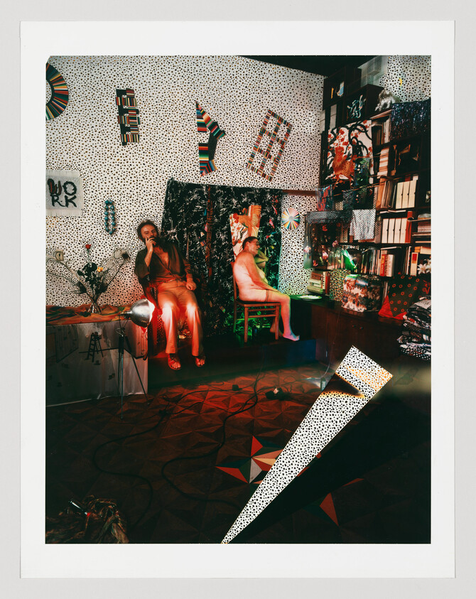 Two people in a cluttered patterned room, one smoking while the other sits naked on a chair.