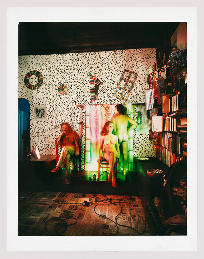 Two nude models pose on a small stage while an older man sits nearby in a cluttered studio.