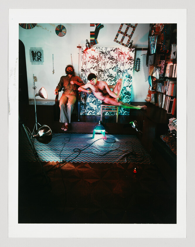 Two men in an art studio, one clothed seated and one nude reclining on a chair amid lights.
