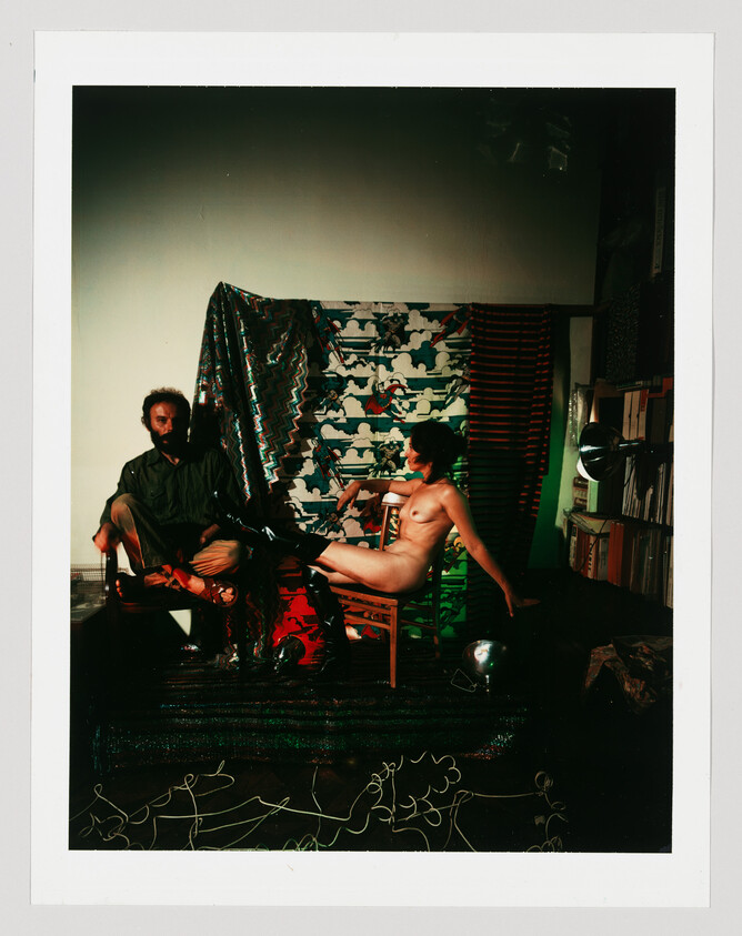A nude woman reclining on a chair beside a seated bearded man against patterned fabric backdrop.