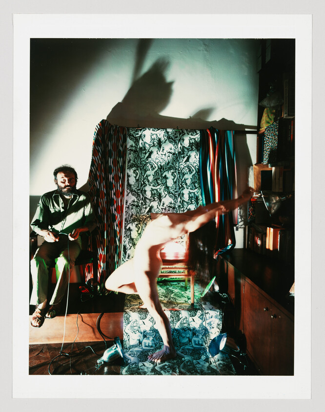 A naked person bends forward on patterned steps while a seated man watches and holds cables.