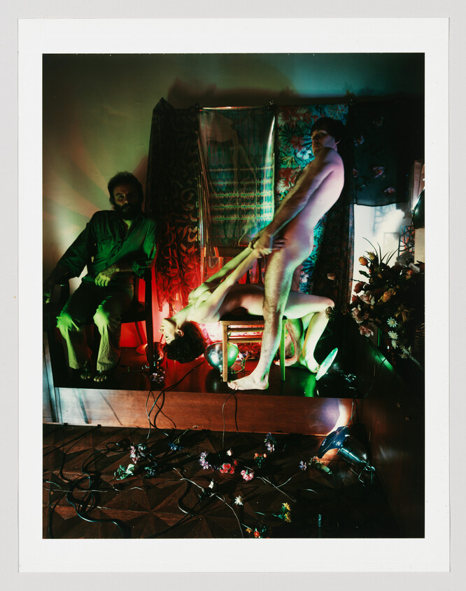 A naked man holds a naked woman upside down while a clothed man watches in a dimly lit room.