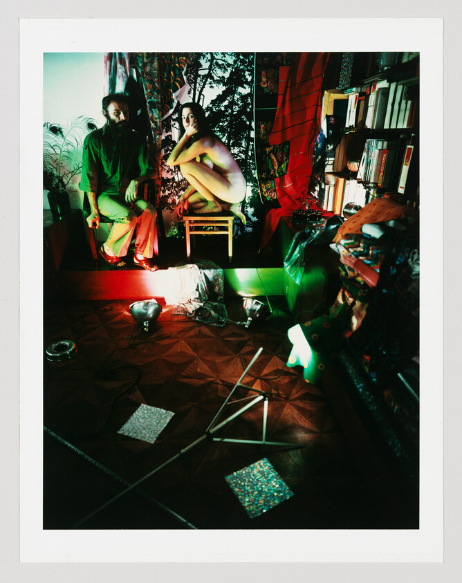 A nude woman crouches on a stool while a bearded man sits nearby in a cluttered, colorfully lit room.