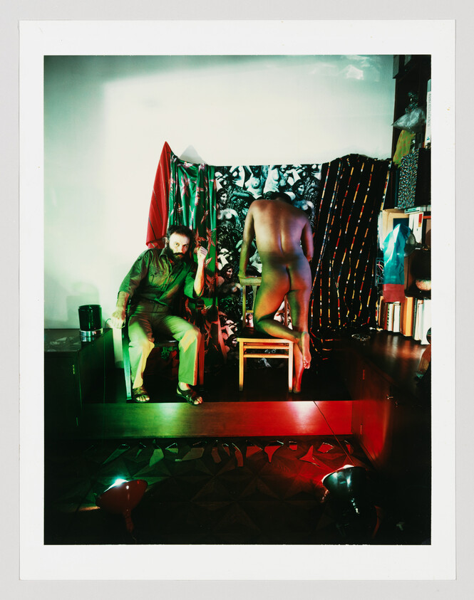 A seated man gestures while a naked person stands on a chair facing away.