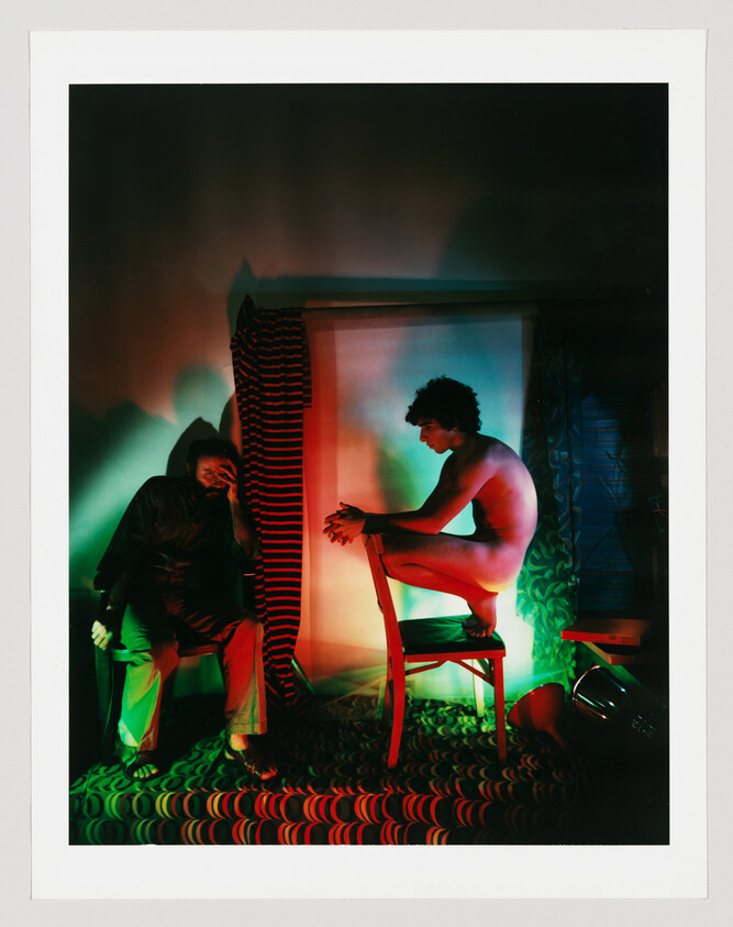 A naked man crouches on a chair facing a seated man who covers his face, under colored lights.