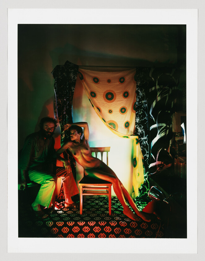 A nude woman poses reclining on a wooden chair while a seated man watches in colorful studio lighting.